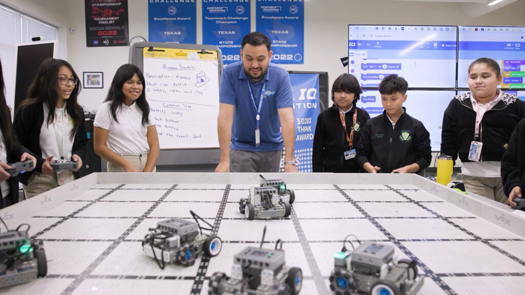 A male teacher in a blue shirt stands between 5 middle school students playing a game with VEX IQ robots.