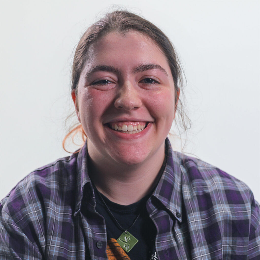 An intern, Lily, smiling. She has brown hair pulled back, a black t-shirt, and a purple checkered shirt.