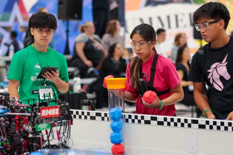 A student in a pink shirt prepares to load during a Push Back match at the 2025 UND Signature Event at the Mall of America. A team mate stands to her left and a volunteer to her right.