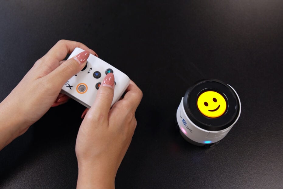 A pair of hands uses a controller to operate a VEX AIM robot displaying a smiling emoji.