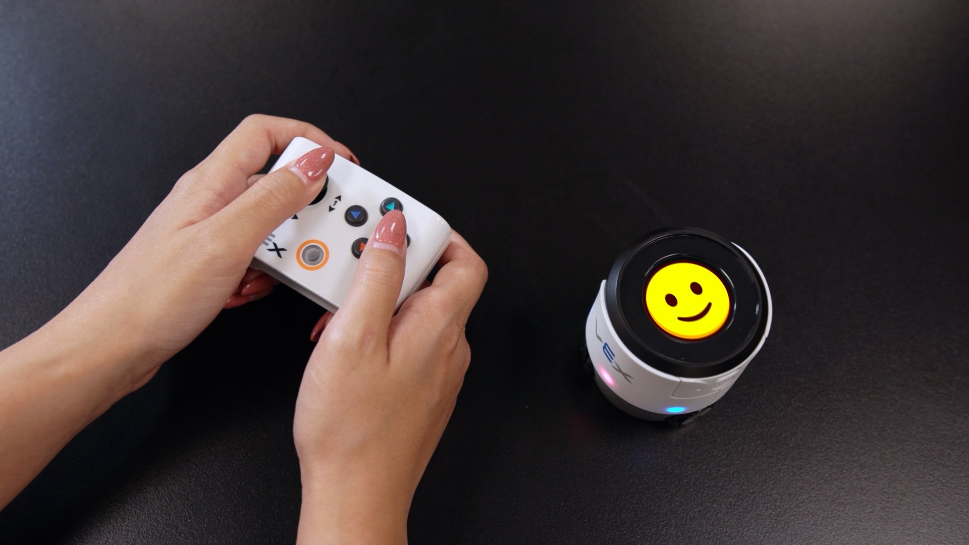 A pair of hands uses a controller to operate a VEX AIM robot displaying a smiling emoji.