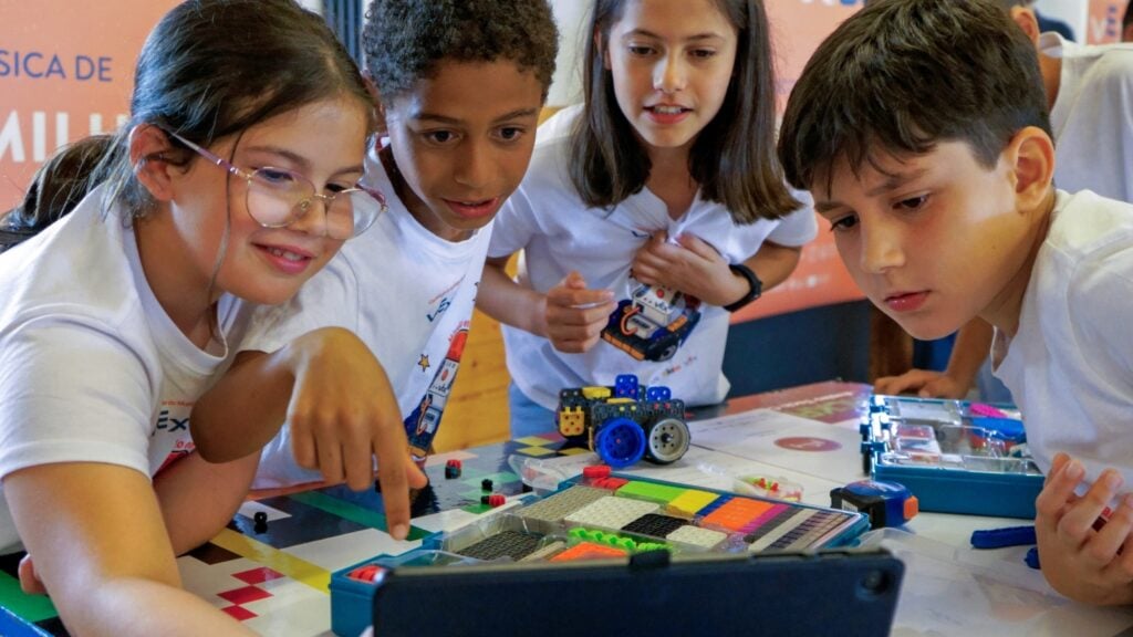 Four students working with VEX GO look at a tablet as the leftmost student taps on the screen facing them. 