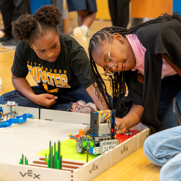 Two students compete in a VEX GO competition. One leans down to adjust something on her robot.