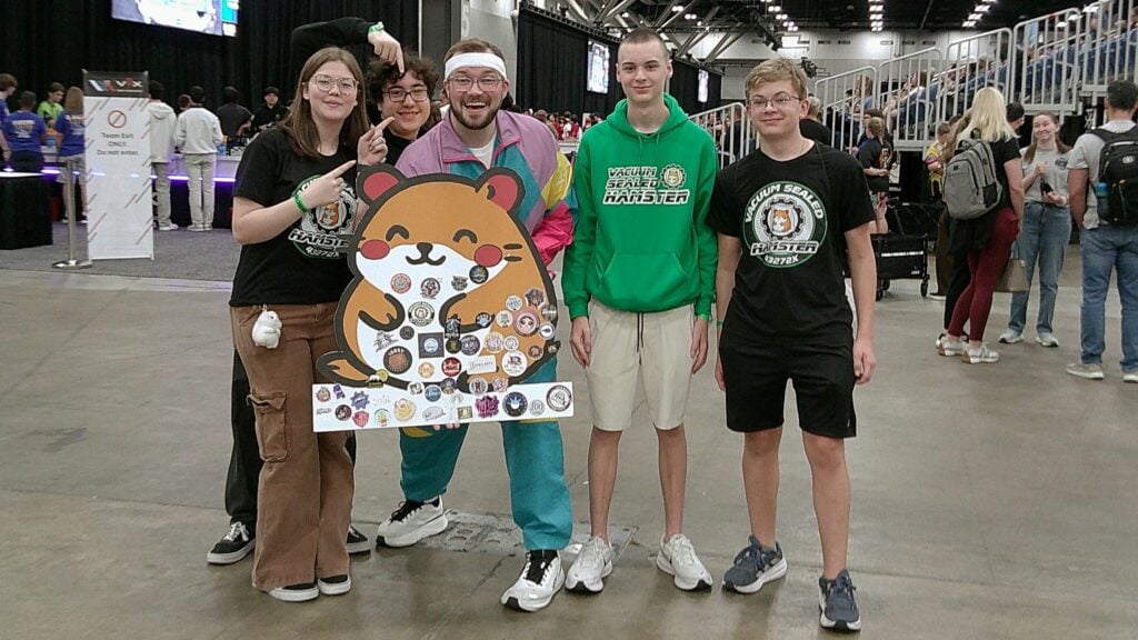 An image by Team 43272X Fox Robotics Vacuum Sealed Hamster of team members posing with Tyler McCrary, Master of Ceremonies, with their team's hamster sign.