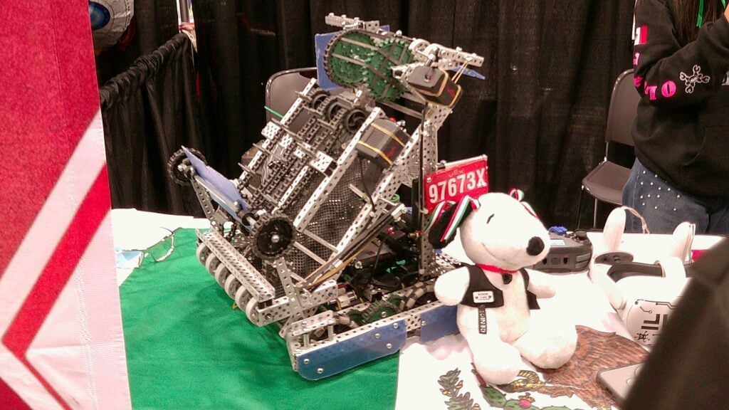 An image by Team 97673X Axolotl of their robot at their booth sitting next to their team's decorated Snoopy plush.