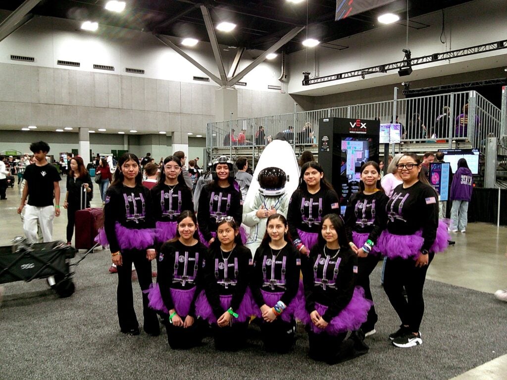 An image by Team 3028G Ladybots of the team posing with their coach in a rocket costume. The girls wear matching purple tutus.